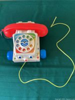 Top view of a vintage Fisher Price Chatter Telephone pull toy showing red handset, rotary dial with numbers, blue wheels, and yellow pull string against green background.