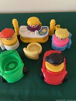 Photo of four Fisher Price Little People chunky figures, three chairs (yellow and blue), two cars (green and red), and a yellow desk.