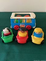 Blue and white mini bus with red wheels and three small cars each carrying a Little People figure including dog in green car, red car with black-haired figure, yellow car with blonde-haired figure.