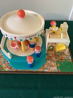 Front view of Fisher Price Little People Merry Go Round showing carousel with canopy, three Little People figures, and adjacent ticket booth with operator figure.