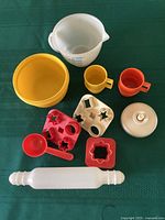 Full lot arrangement on green cloth showcasing vintage Tupperware children's cooking pieces including measuring cup, bowls, cups, cookie cutters, spoon, rolling pin and lid.