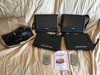 Four Sylvania portable DVD players, two remotes, manuals, and cable bundle displayed on bed