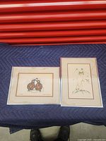 Two framed pictures displayed on a blue quilted blanket on a red backdrop. One horizontal with twin stylized birds, the other vertical abstract animal forms.