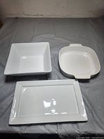 Top view of three white ceramic serving dishes placed on a grey fabric. Includes a square bowl, a rectangular platter, and a square dish with two side handles.