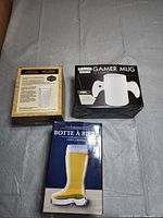Photo of the boxed glass beer stein, Gamer mug, and beer boot from above on grey fabric.