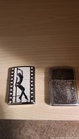 Pair of two Zippo metal lighters on wooden surface, one with black and white film strip graphic, other with silver floral engraving, hinge damaged on engraved lighter.