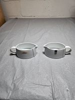 Two white ceramic Denby gravy boats placed side by side on a gray surface, showing front sides with handles and spouts visible.