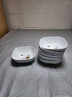 Photo of seven white square Denby soup bowls stacked and one bowl placed beside them on a gray surface against a concrete wall.