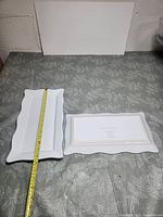 Two white porcelain rectangular serving dishes with wavy edges and gold trim, displayed on a floral patterned cloth background. One dish is longer and narrower than the other. A measuring tape is placed across each dish to show dimensions. The dishes have a glossy finish.