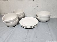 Six white ceramic bowls and five white ceramic plates displayed on a light gray cloth surface against a white wall.