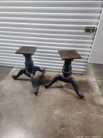 Two large wooden table legs with dark tripod bases and square wooden tops, standing on a concrete floor near a metal garage door.