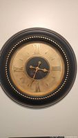 Photo of a modern medium wall clock showing its design and condition: round, black frame with gold accents and Roman numerals on the face.