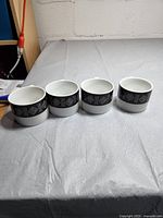 Four white ceramic cups with a black band featuring white geometric symbols, arranged in a row on a white surface.
