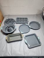 Photo showing seven grey silicone baking pans with light blue and beige silicone handle grips on a grey surface against a white brick background.