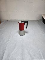 Photo shows red and silver stovetop espresso maker with black handle on a white surface against a white brick background.