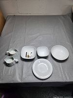 Top-down view of all six white Denby dishes arranged on a grey surface showing two pitchers, two square bowls, one round bowl, and one shallow plate.