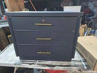 Front view of black wooden storage chest with three drawers and gold handles, closed.