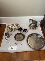 Overview of all 11 pieces showing a variety of silver plated serving ware including pitcher, tray, cup, gravy boat, small dishes, and candlestick holder on a white background.
