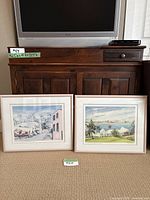 Two framed watercolour prints side by side on carpet in front of furniture and TV. Both prints are similarly framed with beige mats and wooden frames.