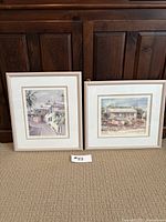 Two framed watercolour paintings signed by C. Holding shown leaning against a wooden cabinet, depicting architectural scenes with trees and flowers, in white mats and light wooden frames.