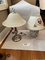 Two authentic table lamps shown side by side on white paper: one with pewter base and cream scalloped fabric shade, the other with ceramic floral base and decorated cream shade with cutout hearts.