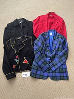 Four women's wool blazers and coats laid flat on beige carpet, showing different patterns and colors: plaid, solid red, black with geometric patterns, black with dots.