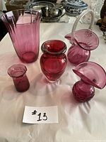 Five cranberry glassware pieces displayed on a table, showing varying shapes and sizes including vases and a basket style piece with handle.
