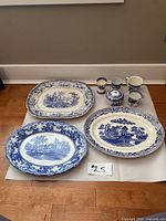 Overview of all eight pieces: three large serving platters with blue and white rural and floral designs, three egg cups, creamer, sugar bowl. One large platter shows repair.