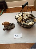 Photo of wire basket filled with assorted beach decor items including shells, stones, starfish and driftwood, alongside a clay fish on a rock.