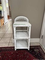 Front view of white painted rattan 3-tier shelf with curved back on top tier, standing on tiled floor and red patterned rug.