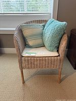 Front view of the wicker armchair with two throw pillows and one seat cushion inside a room with carpet and window.
