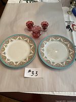 Two antique Royal Worcester dinner plates next to three pink glass candle holders with ruffled edges, all displayed on a table covered with a beige paper surface.