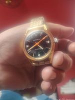 Clear photo of the Caravelle men's watch with black dial, orange second hand, gold-tone case and metal link band, held in hand showing details of dial and case.