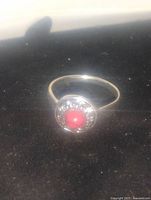 Close-up photo of a gold-tone ring featuring a round red faux gemstone center, surrounded by clear rhinestones, on a thin band.