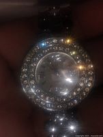 Front view of women's bling watch showing round face with two rows of clear rhinestones surrounding silver dial with Roman numerals and linked metal bracelet.