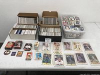 Photo showing multiple cardboard boxes filled with baseball cards, some loose cards and some in protective sleeves laid out on table.