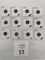 Twenty Indian Head pennies individually housed in white cardboard sleeves with handwritten dates and conditions noted, arranged in rows on a white surface.
