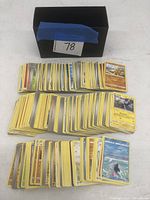 Wide top-down photo showing three stacks of Pokémon cards with a small black storage box behind them with blue tape and the number 78 on a label