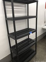 Full view of black plastic 5-tier shelving unit against white wall, showing five perforated shelves and vertical supports.