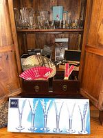 Photo showing a wooden cabinet with various barware including boxed Mikasa shot glasses, hand fans, corkscrews, and an aerating pourer.
