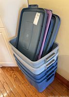 Stack of five plastic storage bins with several lids resting on top, showing blue and gray bins with handles and lids in dark gray and purple.