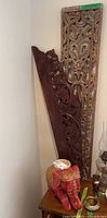 Overview of ceramic red elephant on table with two vertical carved wooden panels leaning against wall behind it.