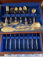 Full view of the wooden case containing the silver-toned flatware with blue velvet lined slots. Shows the organized arrangement of forks, spoons, a butter knife, larger serving spoon, and specialty utensils wrapped in plastic.
