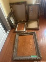 Six vintage-style large art frames arranged on wooden floor showing ornate decorative molding in bronze and gold tones. Frames range in size with two frames missing glass.