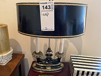 Front view of lamp showing black ceramic base and two candle-like bulbs, black drum shade with gold trim.