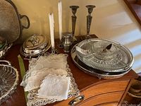Glass lazy Susan, silver serving dishes, candlesticks with candles, and doilies arranged on wooden furniture.