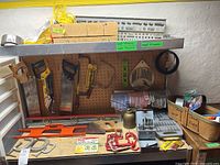 Photo of pegboard wall and workbench with an assortment of hand tools including saws, clamps, levels, a brass oil can, drill bits, gloves, rope, tape and wire.