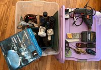 Two plastic totes filled with various tools and household items including bottles, electrical devices, and boxed pathway lights.