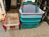 Photo showing three stacked teal plastic storage totes with grey lids and one smaller clear plastic tote with red handles beside them on the floor
