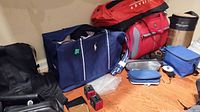 Photo showing a variety of travel bags including a navy Olympia bag, a red insulated lunch bag, multiple wallets, blue toiletry bags, and travel accessories on the floor.